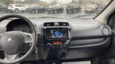 Driver's view from inside a car shows the dashboard and center screen while a row of cars sits in a rainy parking lot outside the windshield, with a few raindrops on the glass and an overcast street scene beyond.