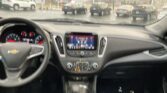 Driver's view of a car interior: steering wheel, center touchscreen, and gear shifter with a rainy parking lot outside through the windshield