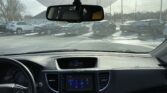 View from the driver’s seat: dashboard and center touchscreen visible, parking lot seen through windshield with several cars and a snowy/icy surface, rearview mirror in frame.