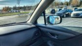 View from inside a car looking through the windshield at a parking lot with many cars and autumn trees in the background, side mirror reflecting more vehicles.