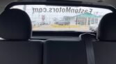 View through a car's rear window of a dealership lot with several parked cars and a showroom building in the background, under overcast sky.