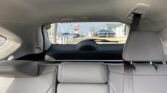 View from the backseat of a beige leather car; rear seatbelt buckled, outside is a dealership lot with cars and signs.