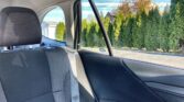 Back seat of a car with a gray patterned headrest and on-seat belt, looking out the rear passenger window at green hedges and a blue sky outside.
