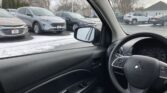 View from the driver’s seat of a car showing the steering wheel and dashboard; outside, a row of parked cars in a snowy dealership lot.