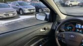 Interior car cabin view looking out at a parking lot with several parked cars and light snow on the ground; dashboard and steering wheel visible on the right.