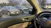 View from the driver’s seat: steering wheel and door panel with a rain-speckled windshield showing a parking lot of cars outside at a dealership/lot.