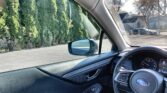 Inside a car cabin showing the steering wheel, dashboard, and driver-side window with a view of trees and houses outside.