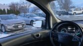 Interior of a car parked in a snowy lot, with the steering wheel and dashboard visible and other parked vehicles outside the windshield.