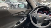 Inside a car on a rainy day, showing the steering wheel and instrument cluster with raindrops on the window and parked cars outside.