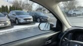 View from inside a car looking through the windshield at a dealership lot with several parked SUVs and sedans, some snow on the ground and in the air outside. The driver’s side mirror is visible in the foreground.