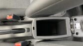 Interior of a car showing the center console with an open storage slot, handbrake lever, and red PRESS seat belt buckles.