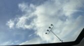 Blue sky with scattered clouds seen through a car sunroof; stadium light poles appear along the right side.