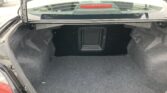 Open car trunk with dark gray carpet, showing a spacious cargo area and a small hatch in the back wall; tail light visible at left edge.