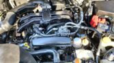 View of car engine bay with battery (red terminal cover), air intake, hoses, and coolant reservoir under the hood.