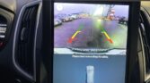 Vehicle rear-view camera display showing the parking lot with several cars and guide lines; warning text reads 'Please check surroundings for safety.'