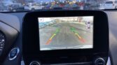Rearview camera display in a car showing parking guidelines (green, yellow, red) over a snowy parking lot, with a top-down car icon on the right side.