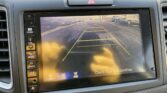 Car infotainment screen showing rearview camera with yellow guidance lines (parking aid) and surrounding cars visible in a parking lot.