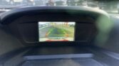 Car dashboard screen showing a rearview camera view with guiding lines, displaying a parking lot with cars and storefronts in the distance.