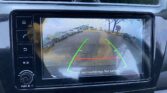 Car infotainment screen showing a rear-view camera: green guiding lines and a red zone, warning to check surroundings for safety as you back up in a parking lot.