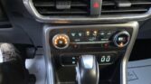 Close-up of a car's center console: climate control panel with orange lit dial, hazard button between vents, and the gear shifter in foreground, plus a 12V port nearby.
