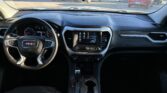 View from the driver’s seat inside a GMC SUV, showing the steering wheel, digital gauge cluster, center touchscreen, and gear shifter with cupholders; parking lot with cars outside the windshield.