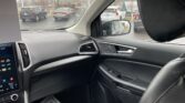 Interior car cabin with black dashboard, touchscreen display, and steering wheel visible; windshield shows a rainy parking lot outside with several parked cars and a pale sky.