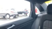Blurred view from inside a car’s back seat, looking out the window at parked cars outside with bright sunlight washing the scene. A headrest and door panel are visible in the foreground.