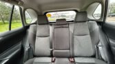 Rear car seats in a dark fabric upholstery with three headrests and visible seat belts, view through rear window to a parking lot.