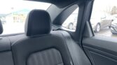 Rear car interior: backseat with black fabric and white stitching, headrest visible, window showing parked cars outside and a blue canopy structure in the distance.