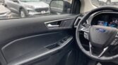 View from the driver’s seat of a car: black dashboard and steering wheel with Ford emblem, looking out at a rainy parking lot with other vehicles.