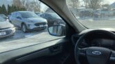Front-seat view from inside a car parked in a snowy lot, with other vehicles visible through the driver's window and the steering wheel in the foreground.