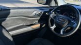Inside a GMC SUV with a right-hand drive steering wheel, dashboard gauges and door controls visible; outside is a sunny parking lot and storefronts.