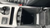 Center console of a car with two cup holders, a small open storage cubby, and a red seat belt buckle nearby on a black leather seat.