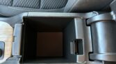 Open car center console storage with dark inner compartment and wood trim on left, showing cup holder area and seat with white stitching nearby