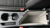 Center console area of a car interior with cup holders, storage compartment, and red seat belt buckles near black seats with white stitching.