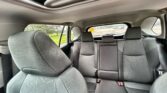 Rear seating of a car interior with gray patterned upholstery and three headrests, seen from the passenger side, with a sunroof above and a glimpse of the outside greenery through the windows.