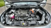Open car hood showing engine bay with battery, pink coolant reservoir, and various hoses and components visible in a gray vehicle.