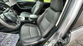 Interior of a car front seats and steering wheel viewed from open driver's door; gray fabric seats with geometric stitching, center console, and cupholders; green park visible outside