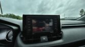 Car dashboard with center touchscreen displaying radio, vents, and hazard button; cloudy sky visible through windshield.