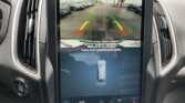 Rearview camera display showing a parking lot with vehicles on both sides and green/yellow/red guide lines for parking assist.