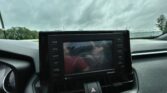 Car dashboard touchscreen showing rear-view camera guidelines over a road, with cloudy sky outside the windshield,