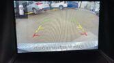 Rear‑view camera display showing a parking lot with colored guide lines (green, yellow, red) and the caption 'Check surroundings for safety'.