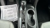 Center console of a car showing an automatic shifter, cup holders, and a paper floor mat beside the driver seat near the pedals.