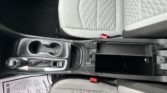 Center console area of a car showing the gear shifter, two cup holders, and open storage compartment beside quilted gray seats with red seat belt latch nearby.