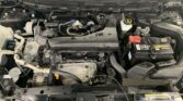Front view of a car's engine bay showing the battery on the passenger side, coolant reservoir, air intake, and various hoses and belts under the hood (Nissan badge on engine cover).