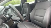 View of the car's front interior: driver and passenger seats in dark gray fabric, steering wheel, center console, and headrests; outside greenery visible through the windows.