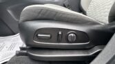 Driver seat adjustment controls on the side of the seat, including a horizontal switch, a round knob, and a secondary lever, with fabric upholstery and a visible seat belt buckle in the background.