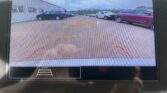 Rear‑view camera display with orange guiding lines showing a parking path, cars parked along both sides, and a light industrial building on the left.