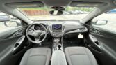 Front interior of a Chevrolet car: steering wheel, center touchscreen, and front seats viewed from the back seat in a parking lot outside the windshield.