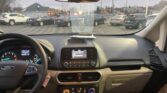 Interior of a car cockpit viewed from the driver's seat, showing a Ford steering wheel, center console, and rearview mirror; outside, a busy parking lot with many parked cars.